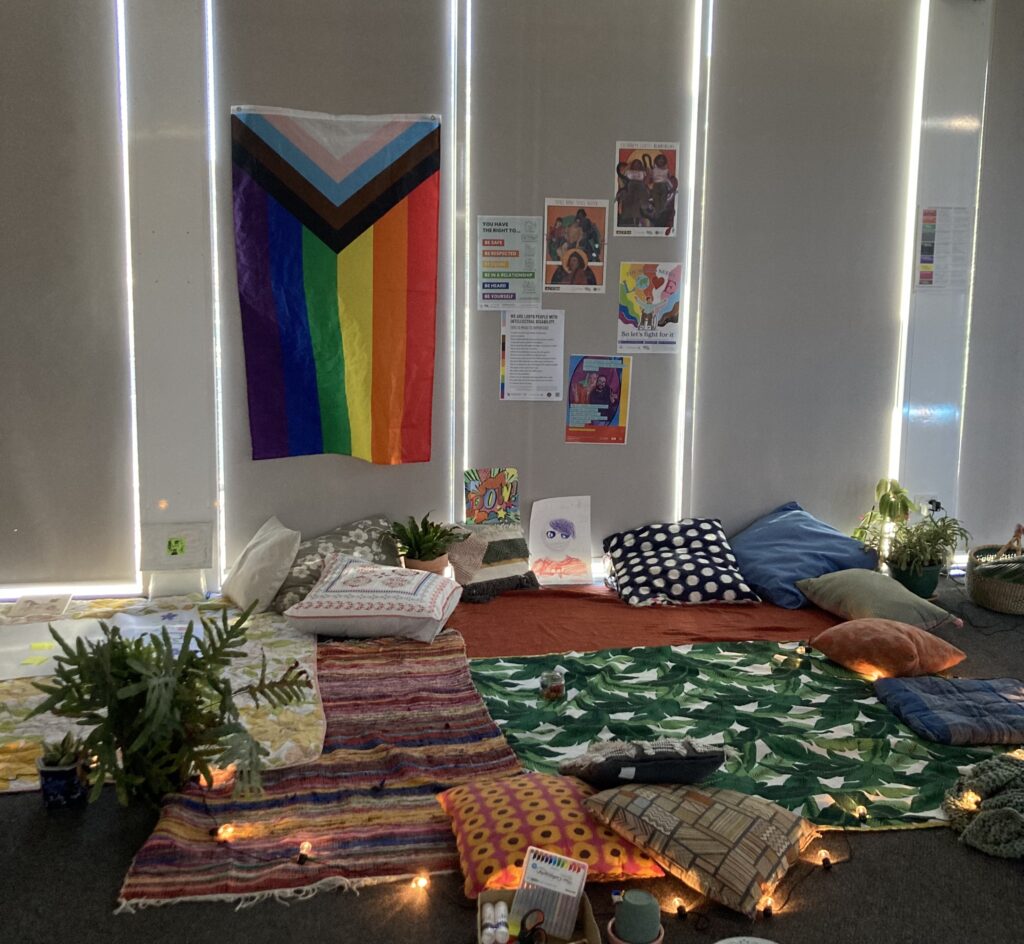 The FUSE space — it is softly lit with fairy lights, filled with cushions and rugs, with a huge progress pride flag hanging on the wall behind along with posters from the Zoe Belle Gender Collective